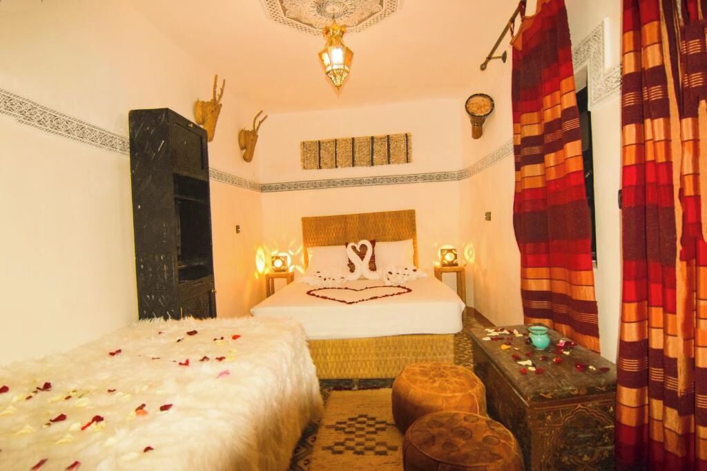 A room with a bed, a wooden chest, a small table, and a wall-mounted shelf.