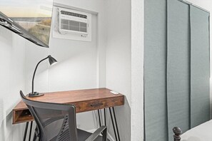 Ein kleiner Büroraum mit Schreibtisch, Stuhl und Stehlampe. Über einem Fenster ist eine Klimaanlage angebracht.