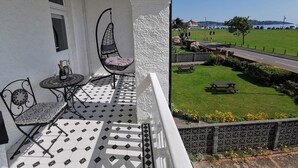 Ein Balkon mit einer Hängematte, einem Tisch mit Flasche und Gläsern sowie Blick auf eine Rasenfläche mit einer Straße und geparkten Fahrzeugen.