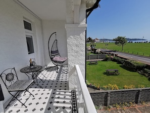Ein Balkon mit einer Hängematte, einem Tisch mit Flasche und Gläsern sowie Blick auf eine Rasenfläche mit einer Straße und geparkten Fahrzeugen.