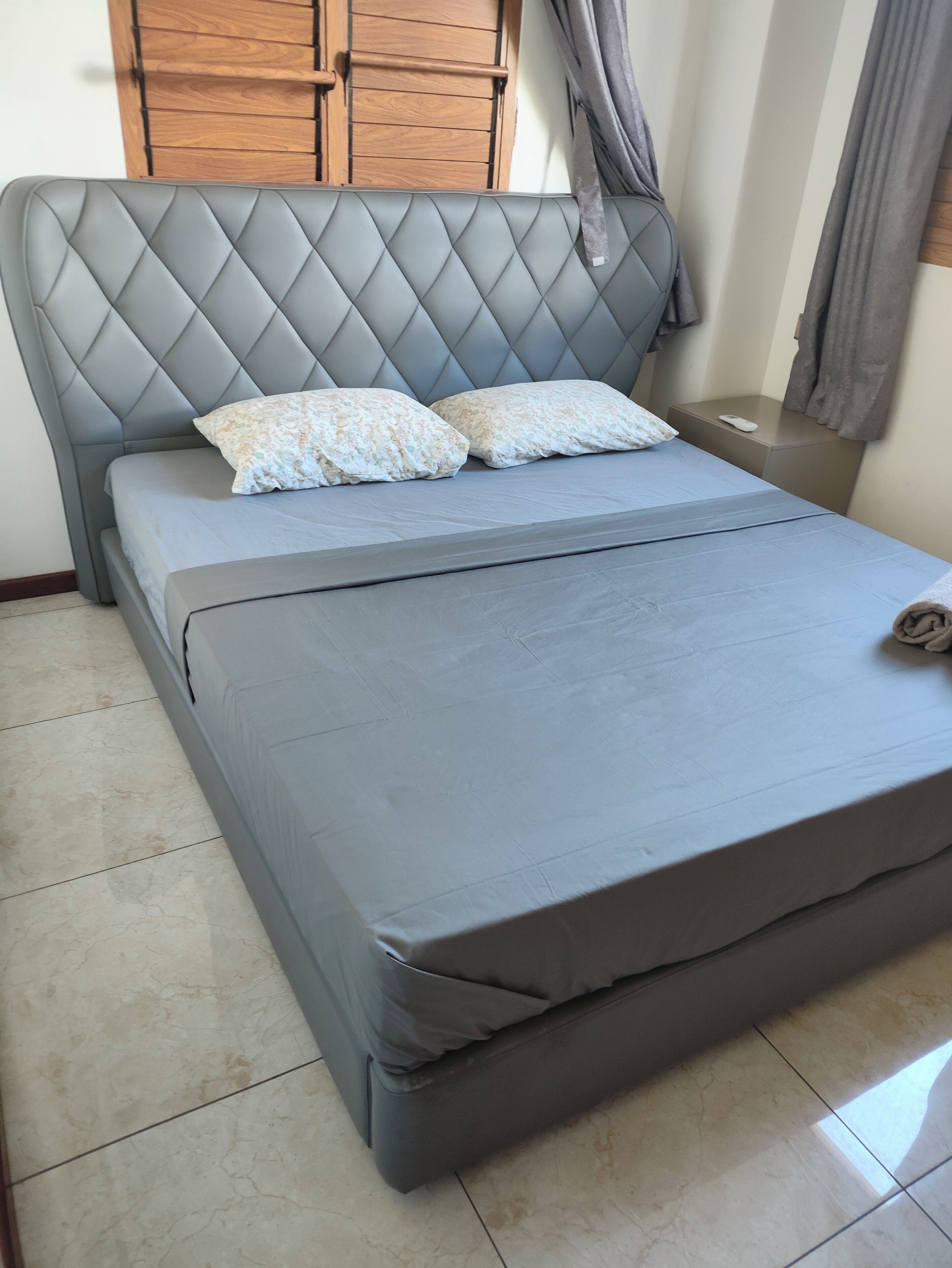 A bed with a grey headboard and two pillows, a bedside table with a remote control, and a window with curtains.