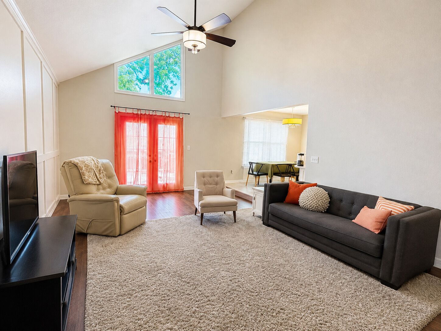 A living room with a flat-screen TV, two leather chairs, a dark sofa with decorative pillows, a ceiling fan, and a dining area with a table and chairs.