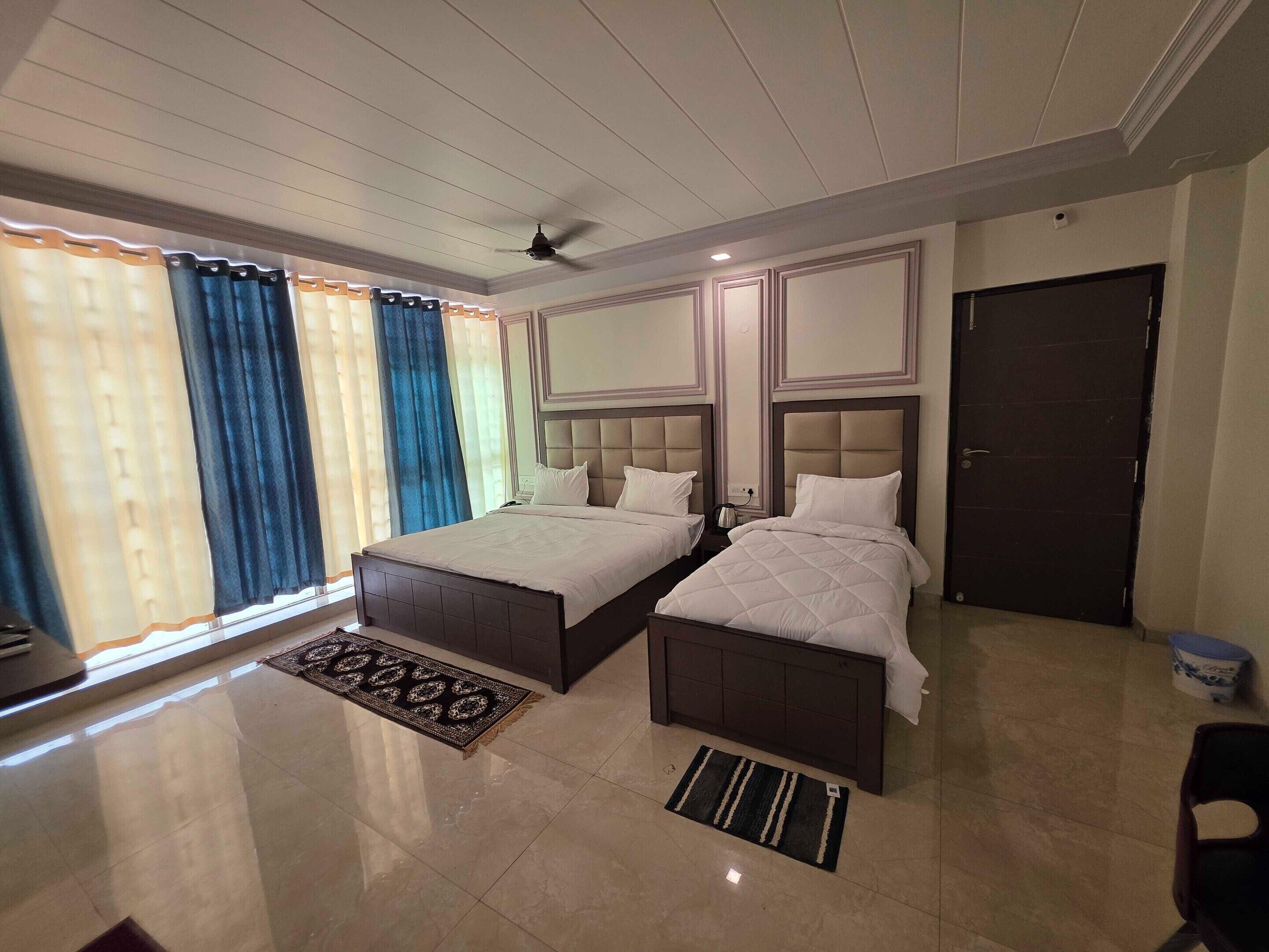 A hotel room with two beds, a ceiling fan, a large window with curtains, and a door.