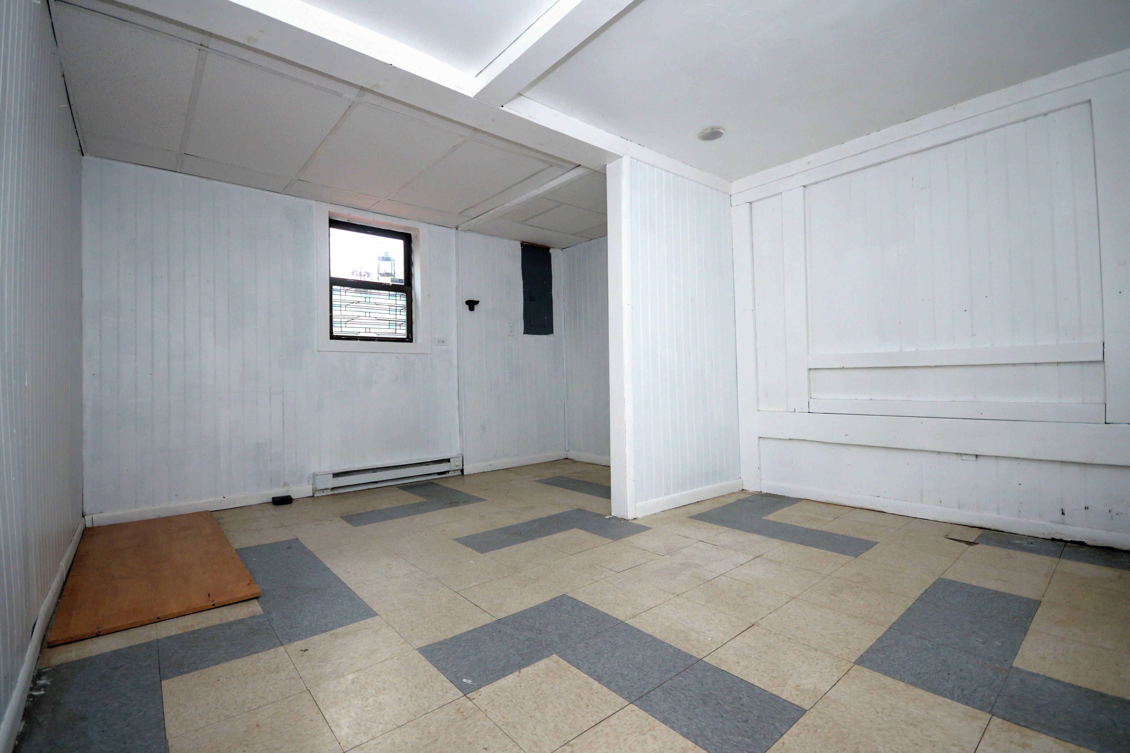 A room with white walls, a window with bars, a door, and a tiled floor with a chevron pattern.