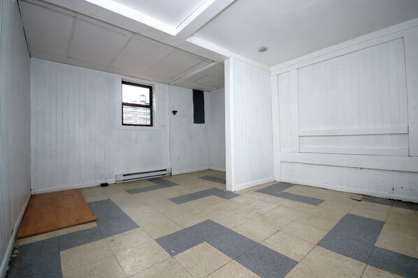 A room with white walls, a window with bars, a door, and a tiled floor with a chevron pattern.