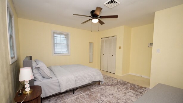 A bedroom with a bed, a bedside table with a lamp, a ceiling fan, a window with blinds, and a closet.