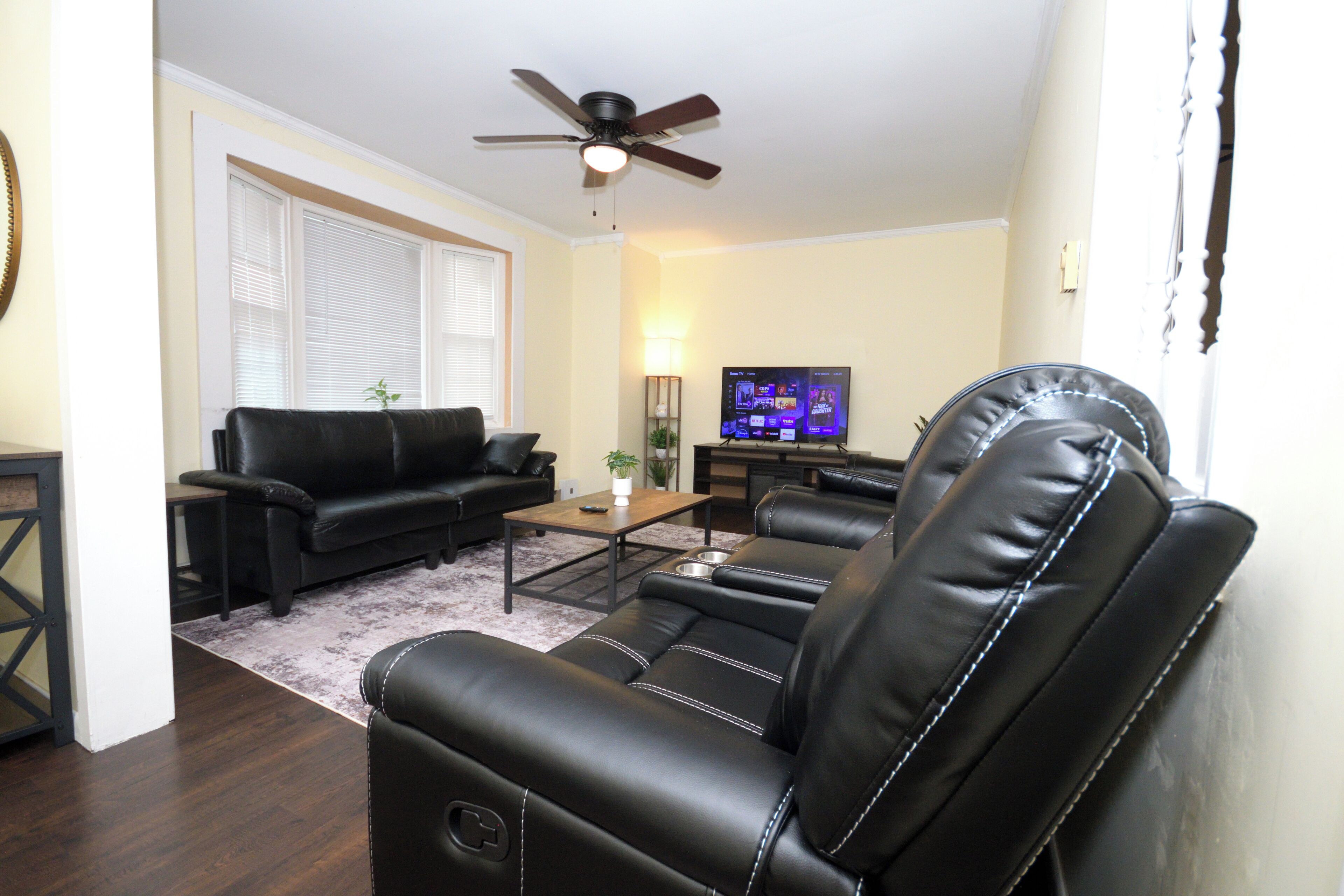 A living room with a black leather sofa, a coffee table, a television, and a ceiling fan.