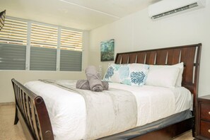 Un dormitorio con cama de madera, ropa de cama blanca, una ventana con persianas y un equipo de aire acondicionado.