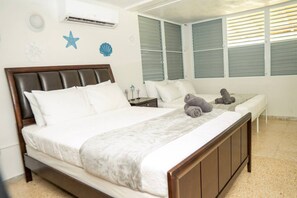 A bedroom with a large bed, a bedside table, a nightstand, and a window with blinds. - Casa Julia Tropical Beach Stay (Luquillo)