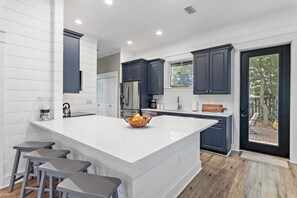 A modern kitchen with a large island, dark blue cabinets, and a view to the outdoors. - 🚲Willow & Waves | Golf Cart, 4 Bikes, Pool, Near Beach🏊 (Inlet Beach)