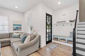 A living room with a beige sofa, a painting of a beach, a black door, and a staircase. - 🚲Willow & Waves | Golf Cart, 4 Bikes, Pool, Near Beach🏊 (Inlet Beach)
