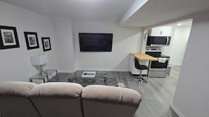 A modern living room with a sofa, a coffee table, and a TV. There is a kitchen area with appliances and a dining table.