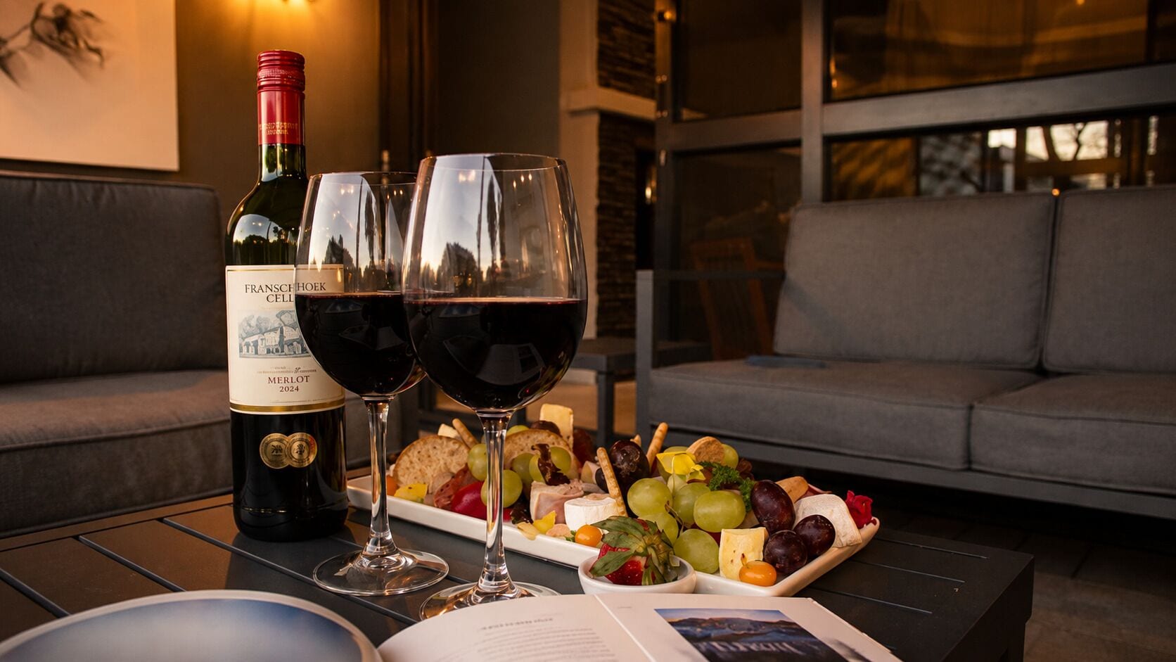 A bottle of Merlot wine, two wine glasses, and a charcuterie board on a table.