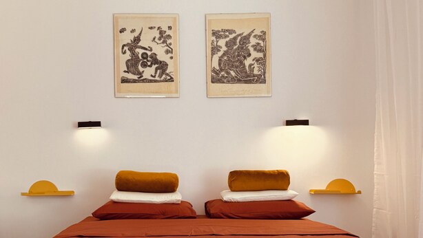 Una habitació amb un llit, dues làmpades penjades a la paret i dues obres d'art amb marc.
