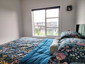 Ein Schlafzimmer mit großem Fenster, einem Bett mit floralem Bettzeug und Blick auf Gebäude.