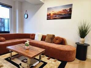 A modern living room with a brown sectional sofa, a wooden coffee table with a potted plant, a black side table with a tall plant, and a framed picture of a pier on the wall. - The Edmonton Nest (Edmonton)