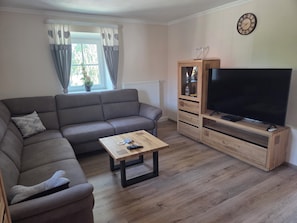 Ein Wohnzimmer mit einer Couch, einem Couchtisch, einem Fernseher auf einem Holzgestell und einem Fenster mit Vorhängen.