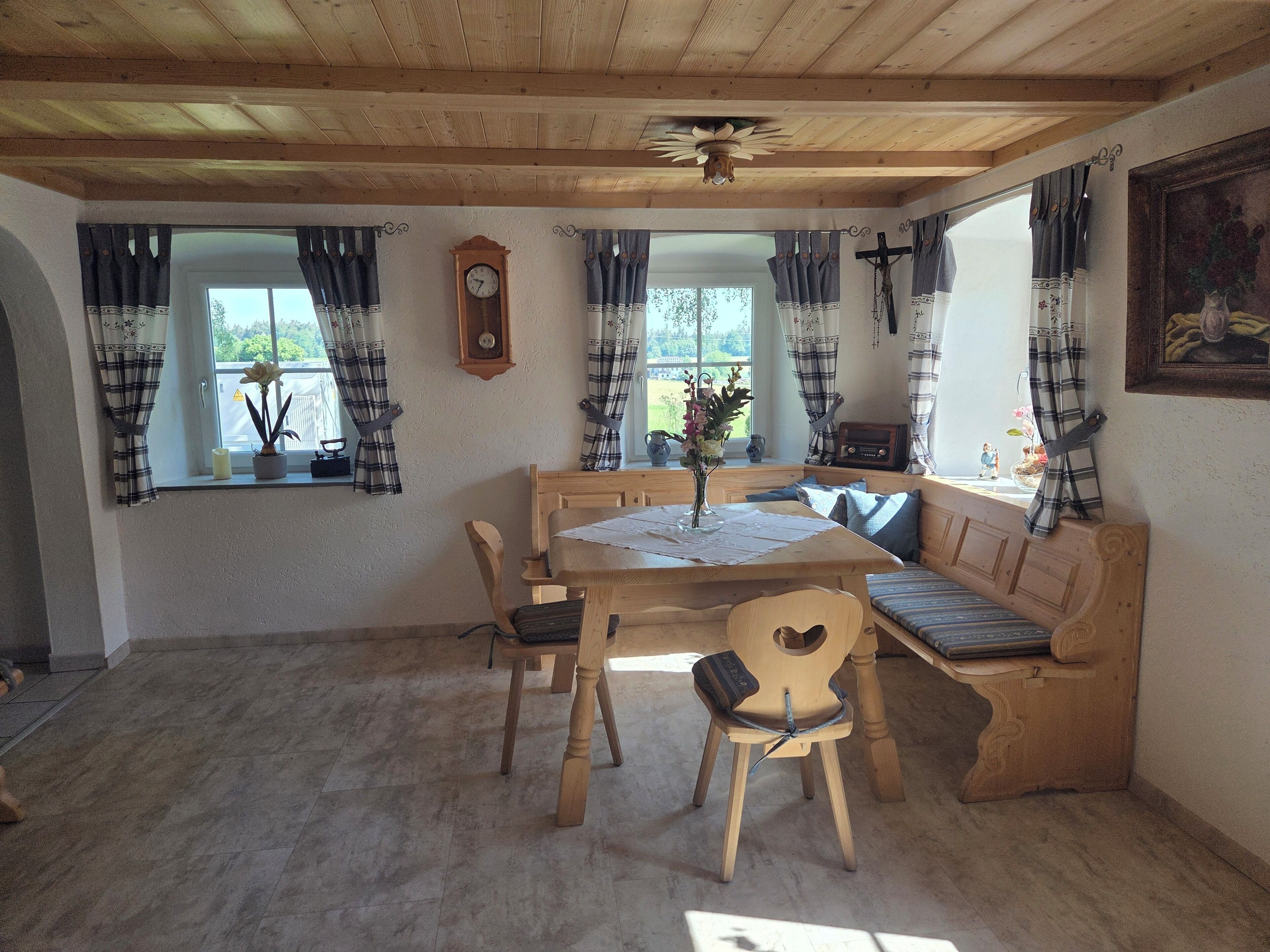 Et spiseområde i træ med et bord, stole og en bænk. Der er et ur på væggen og et vindue med gardiner.