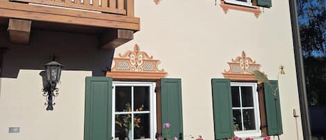 A building with a balcony, multiple windows with shutters, and a flower bed with pink flowers.