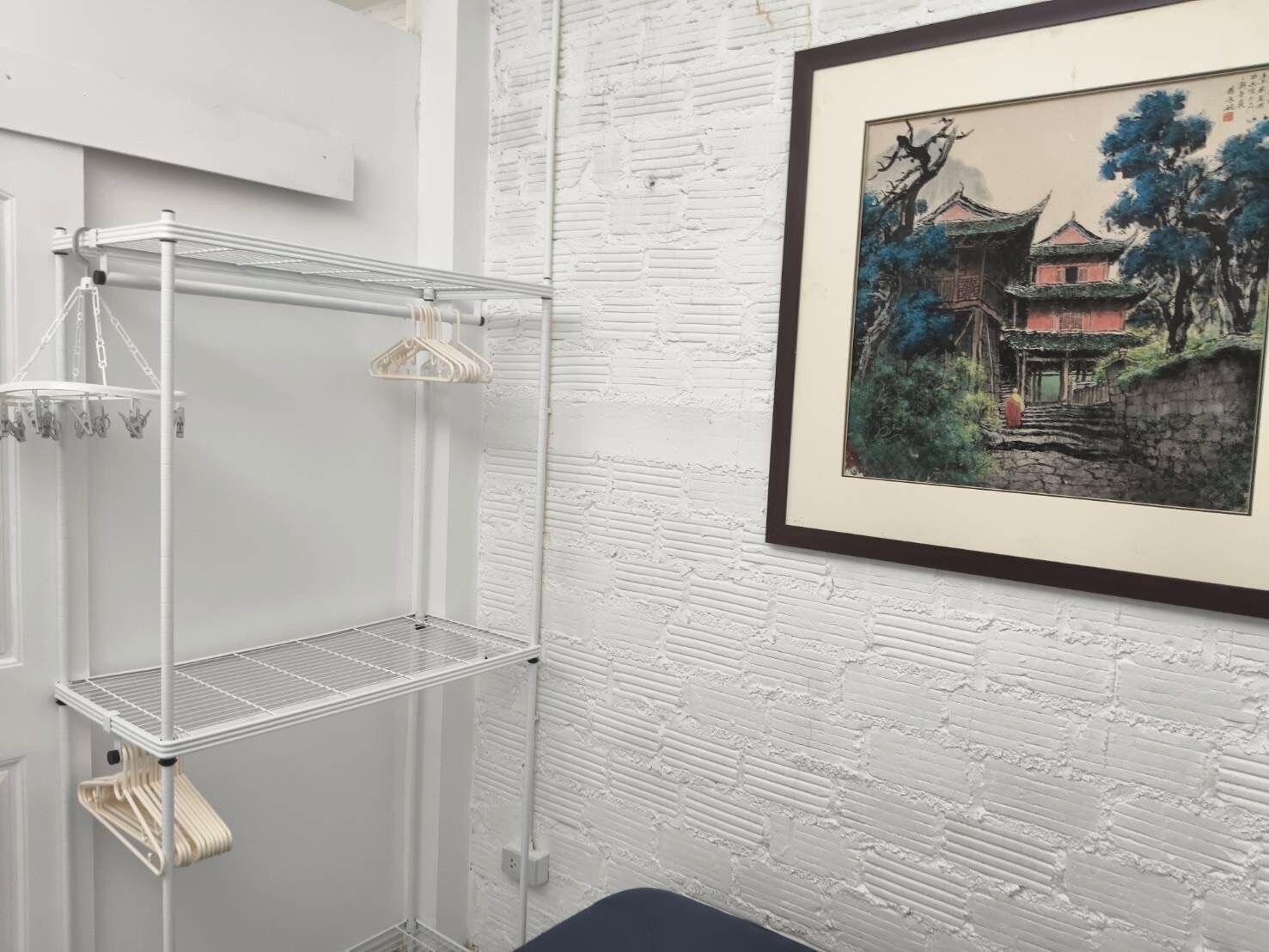 A room with a white brick wall, a metal shelving unit with clothes, and a framed painting of a traditional building.