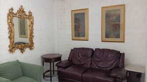 A room with a leather sofa, a green armchair, a round wooden side table, and two framed paintings on the wall.