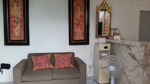A room with a sofa, two pillows, a wall-mounted mirror, and a standing fan.