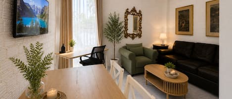 A living room with a wooden dining table, a sofa, a TV mounted on the wall, and a window with curtains.