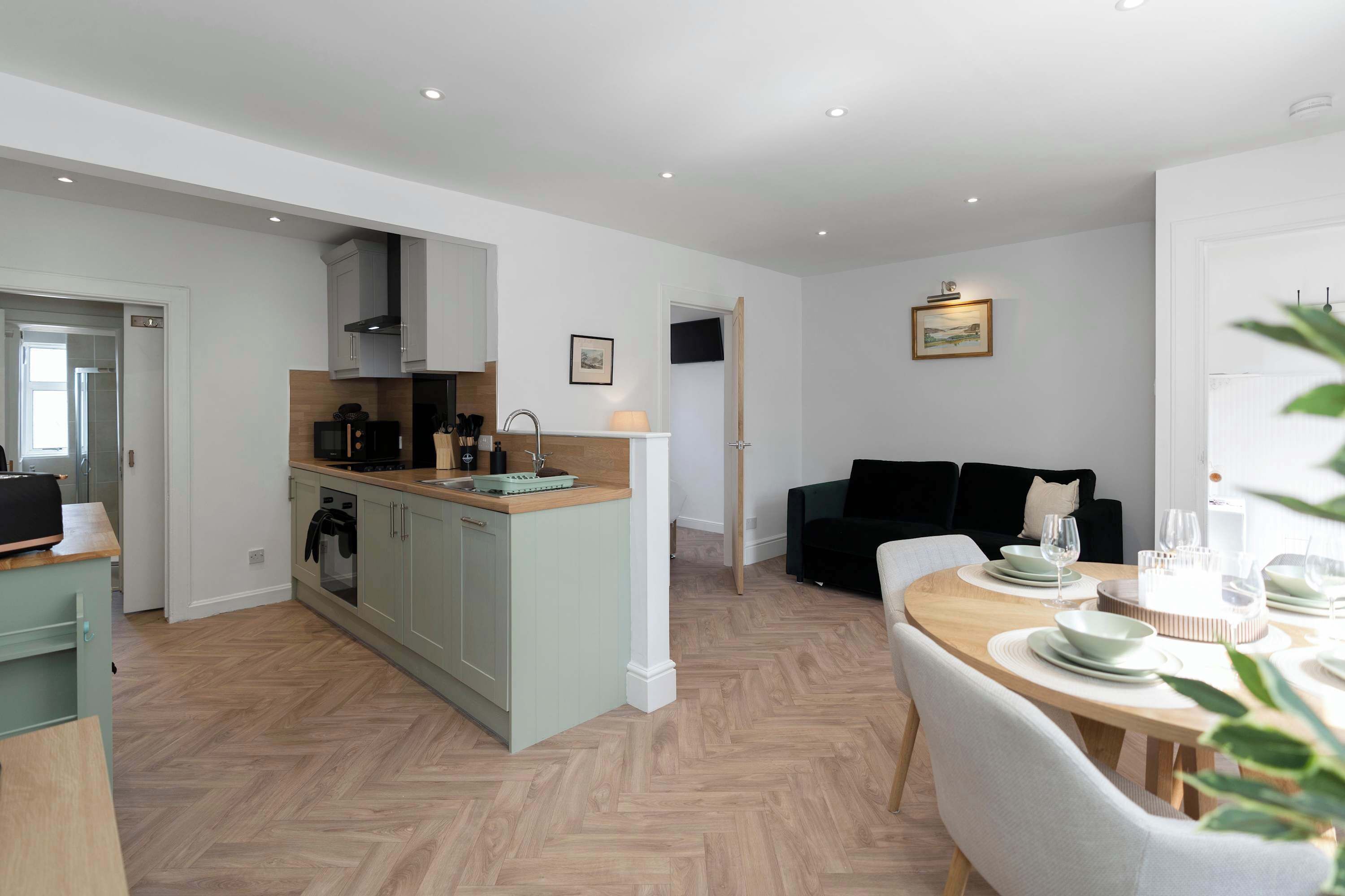 A modern kitchen with a central island, a dining area with a round table, and a black sofa in a living room.