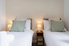 A hotel room with two beds, each with a pillow and a bedside lamp. There is a nightstand between the beds with a lamp and a small drawer. - 1BR Selkirk Town Centre Flat, Parking, Sleeps 4 (Scottish Borders)