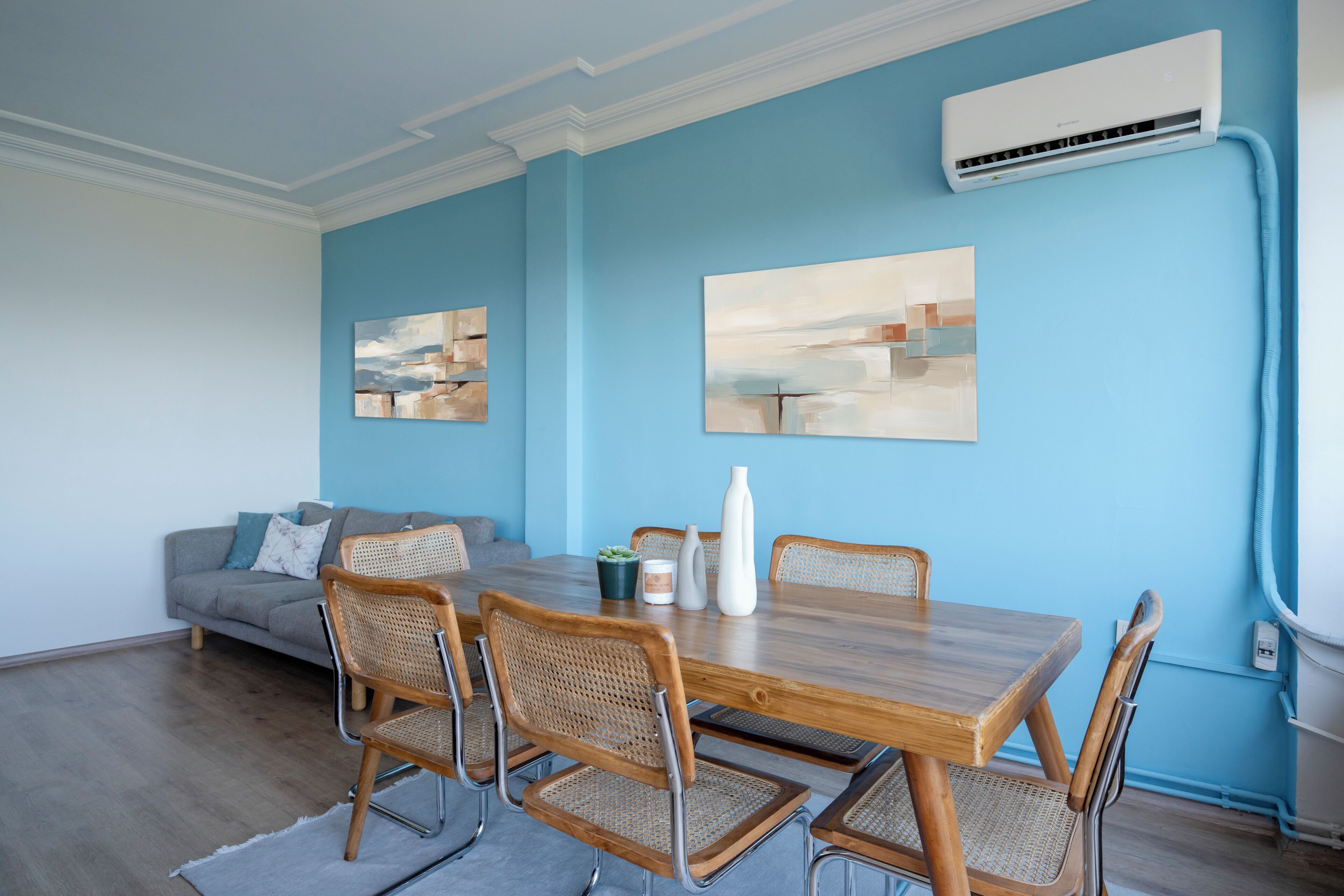 Un coin repas comprenant une table en bois, quatre chaises en osier, un canapé gris et deux tableaux abstraits accrochés au mur.