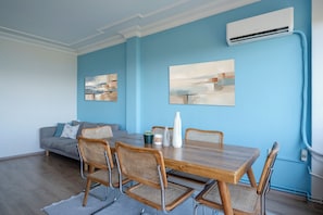 A dining area with a wooden table, four wicker chairs, a grey sofa, and two abstract paintings on the wall. - Bosphorus View with Balcony - 3 Bedrooms 2 Bathrooms (İstanbul)