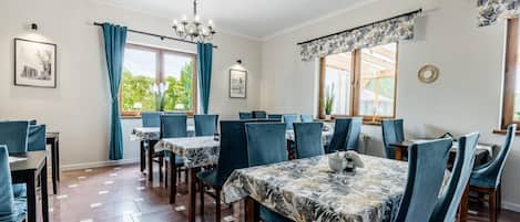 A dining area with multiple tables and chairs, large windows with curtains, and artwork on the walls.