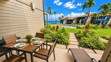 Eine sonnige Terrasse mit Tisch für zwei Personen, umgeben von üppiger Vegetation und Blick auf Pool und Meer.