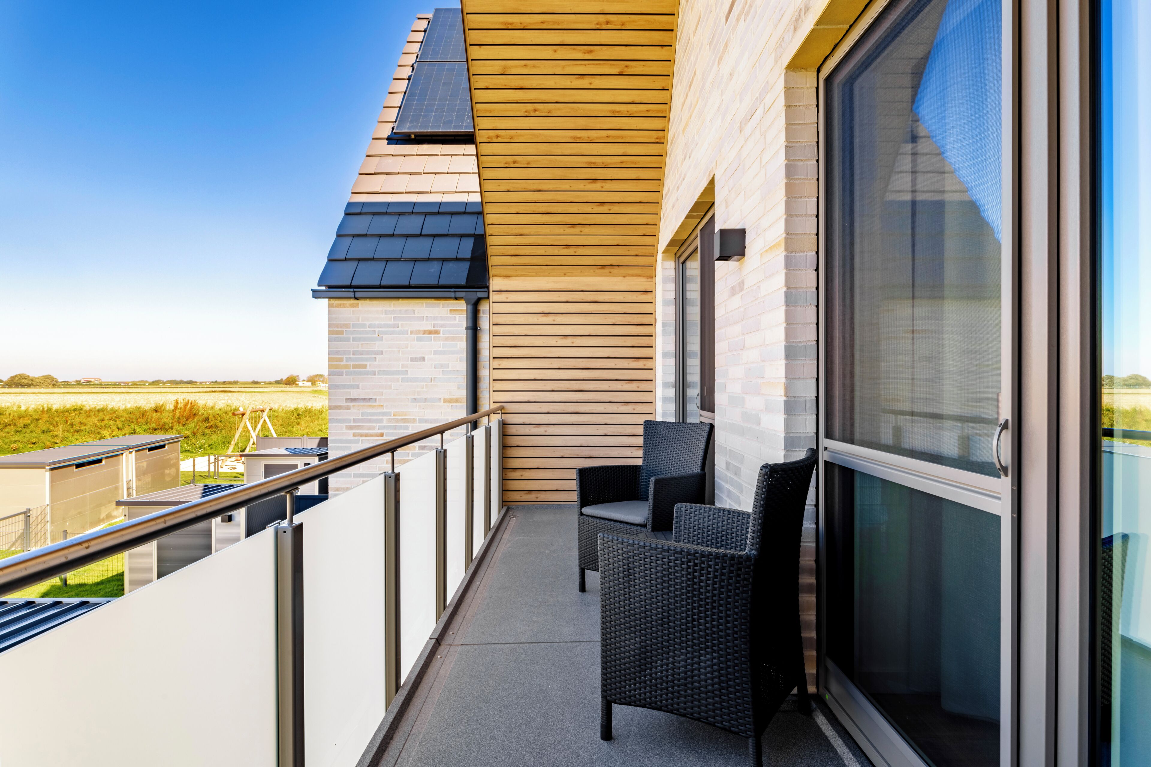 A balcony with a view of the countryside, featuring a wooden slatted ceiling, a glass door, and two wicker chairs.