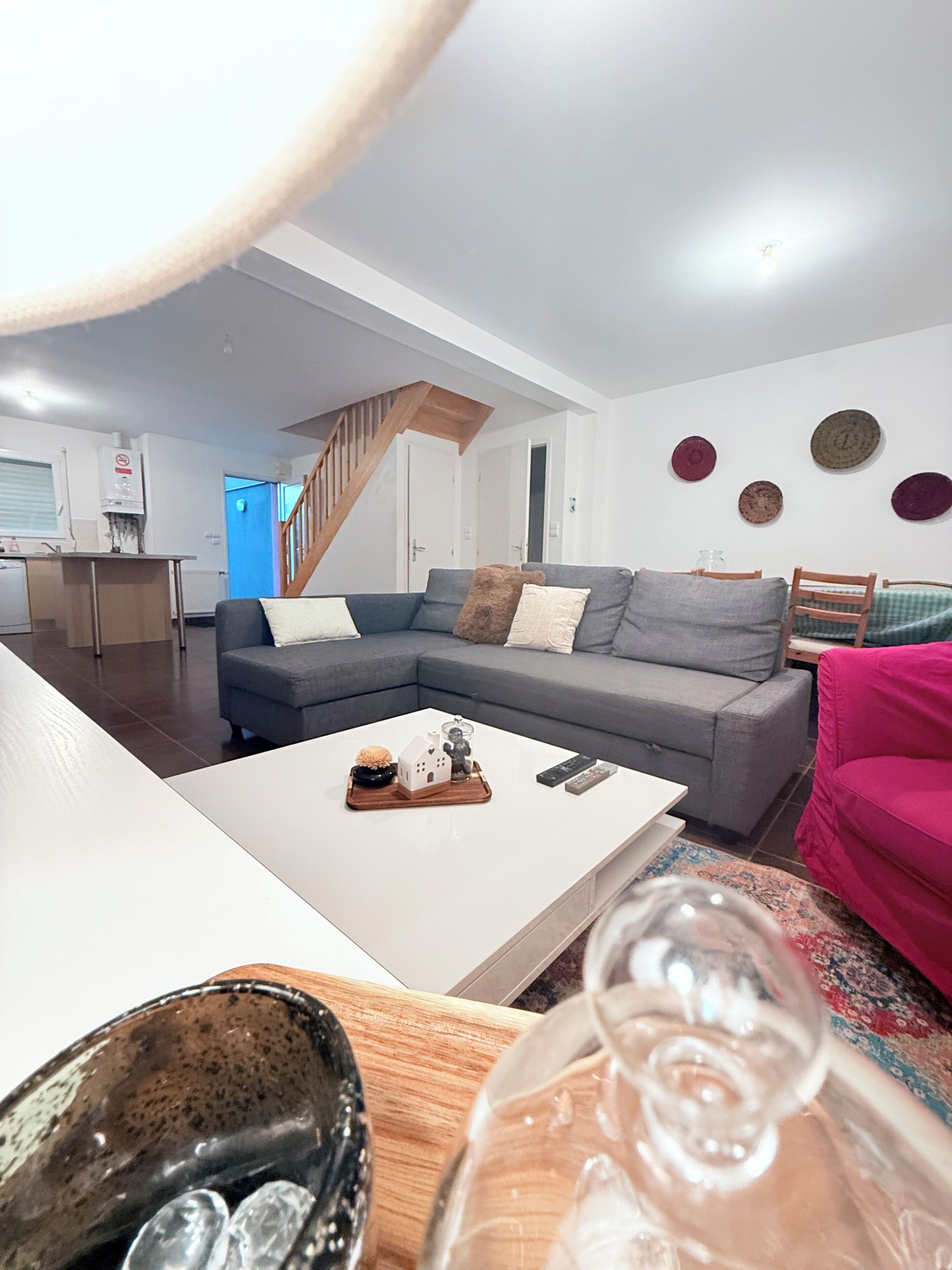 Una sala de estar moderna con un sofá seccional gris, una mesa de centro blanca y un sillón rosa. Se ve una escalera de madera al fondo.