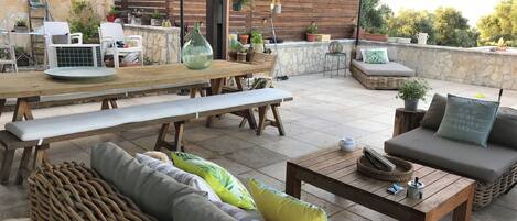 An outdoor patio area with a wooden table, benches, a wicker sofa, and a cushioned seating area.