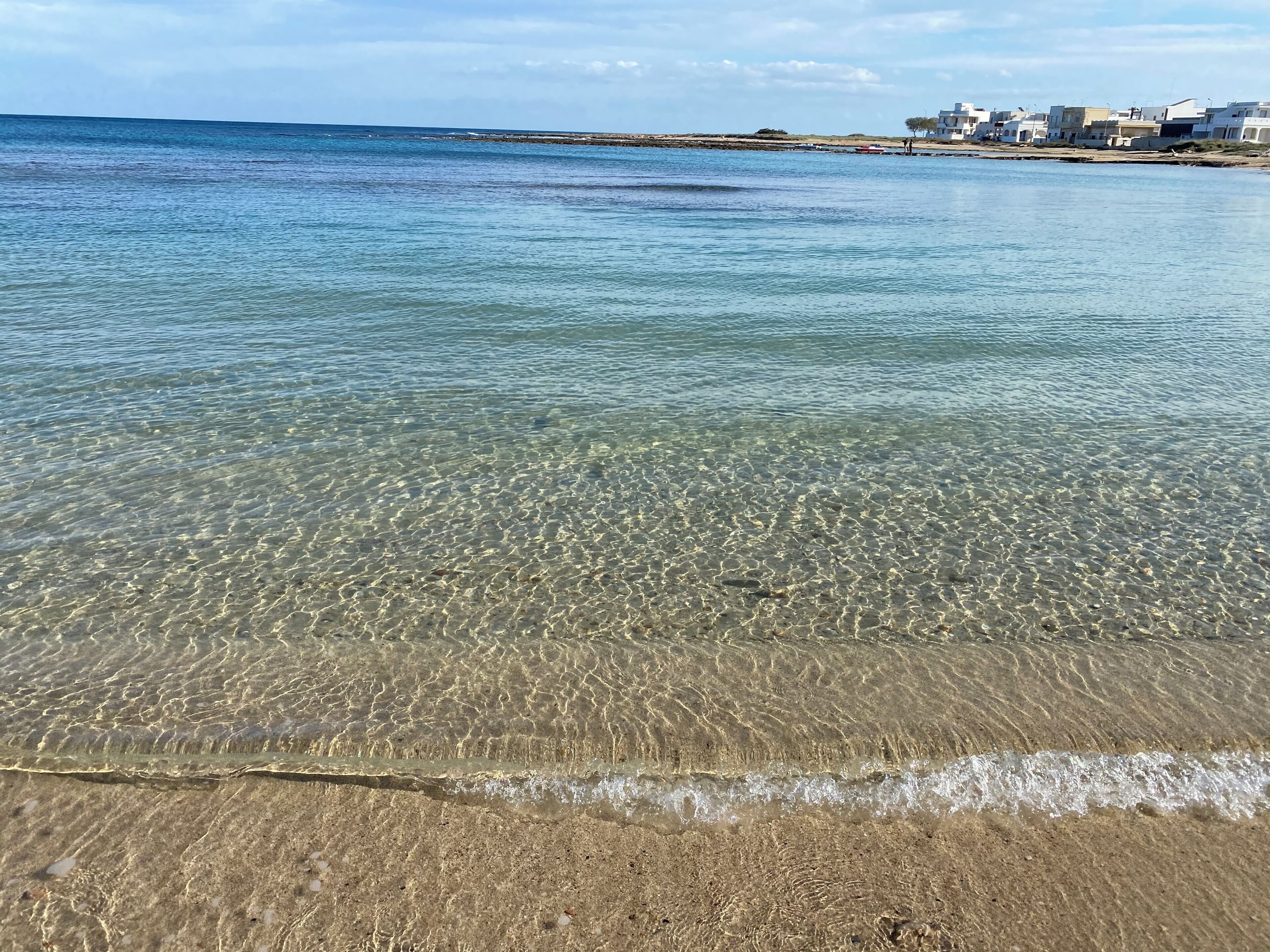 Una playa con aguas poco profundas y cristalinas, y una costa lejana con edificios.