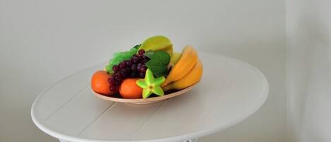 Uma pequena mesa redonda com uma fruteira em cima, duas cadeiras de madeira com almofadas, e um chão branco.