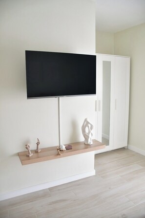 Ein Zimmer mit flachbildfernseher an der Wand, einem weißen Kleiderschrank, einem Holzbodenregal mit Dekorationen und hellem Bodenbelag.