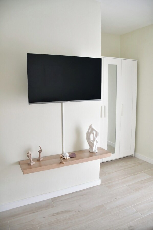 Een kamer met een wandgemonteerde flatscreen televisie, een witte garderobe, een houten plankje met decoratieve objecten en lichtgekleurde vloer.
