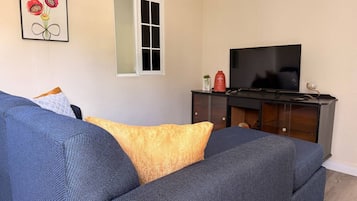 Een woonkamer met een grote bank, een televisie op een standaard en een raam met zonwering.