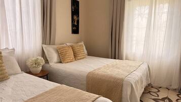 Une chambre d’hôtel avec deux lits, une literie de couleur beige, une petite table avec un vase et des rideaux légers.