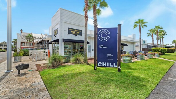 Un edificio moderno con un letrero de Nectar Coffee & Wine, un letrero de Chill Play y un césped bien cuidado con palmeras.
