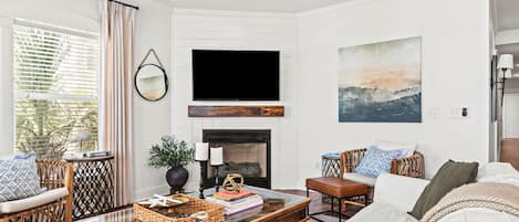 A living room with a fireplace, a flat-screen TV mounted above it, wicker furniture, and a large painting on the wall.