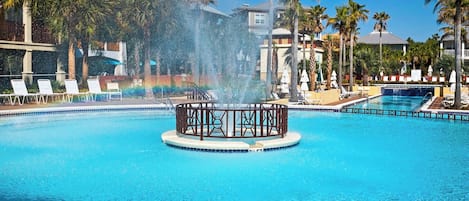 A swimming pool with a central fountain, surrounded by palm trees and lounge chairs.
