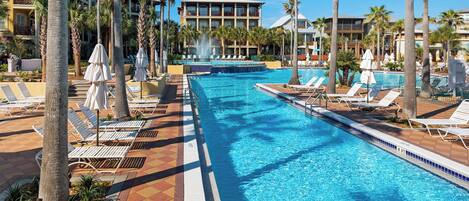 A swimming pool with lounge chairs and umbrellas, surrounded by palm trees and hotel buildings.