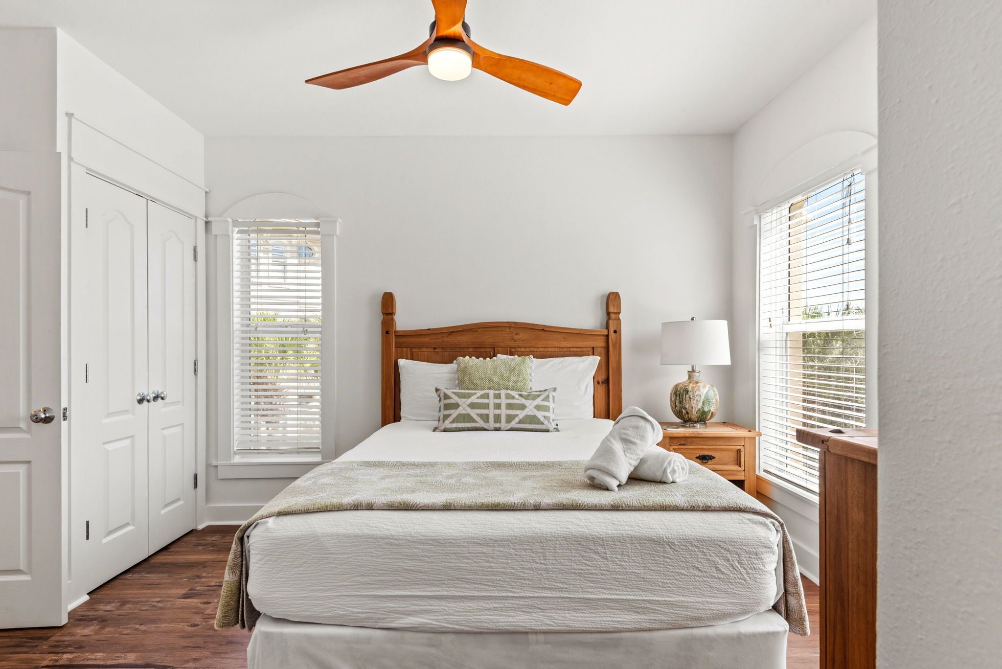 A bedroom with a bed, a ceiling fan, a nightstand with a lamp, and a window with blinds.
