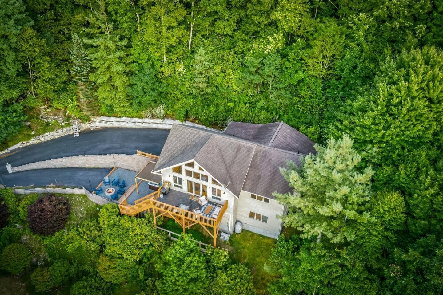 An aerial view of a house surrounded by dense forest, with a visible driveway and outdoor seating area.