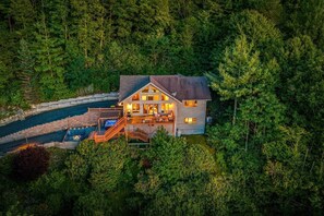 A large house with a deck and a hot tub, surrounded by dense forest.