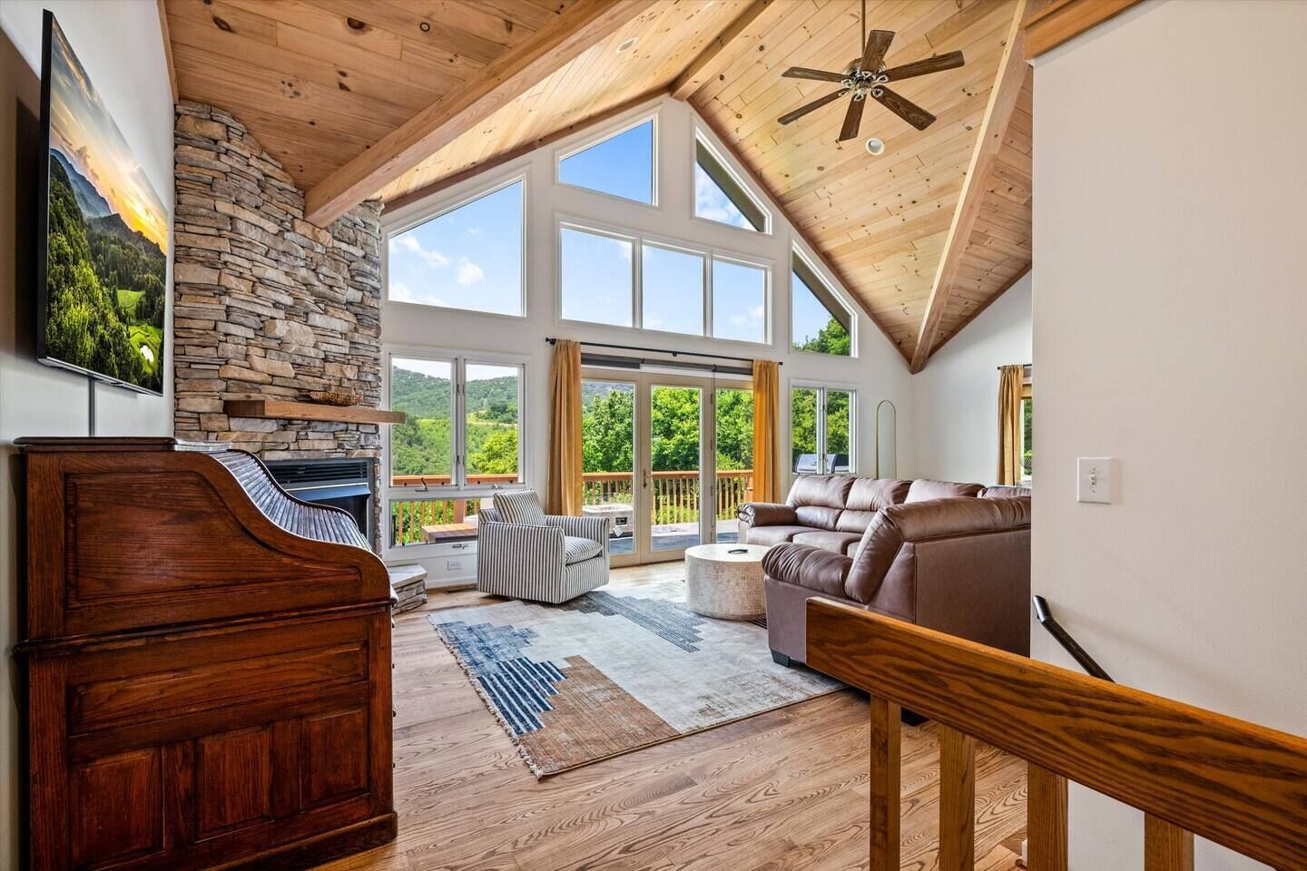 Amplia sala de estar con chimenea de piedra, ventanas grandes, ventilador de techo y escalera de madera.